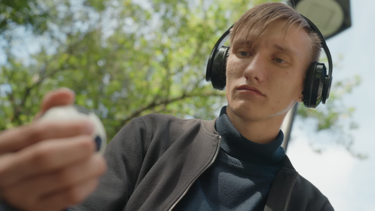 Giovane uomo bianco all'aperto che controlla il telefono e l'auricolare, ritratto candido dal basso di un creatore di contenuti che rivede l'audio mentre indossa le cuffie in un parco alberato, reazione sottile, telefono portatile visibile