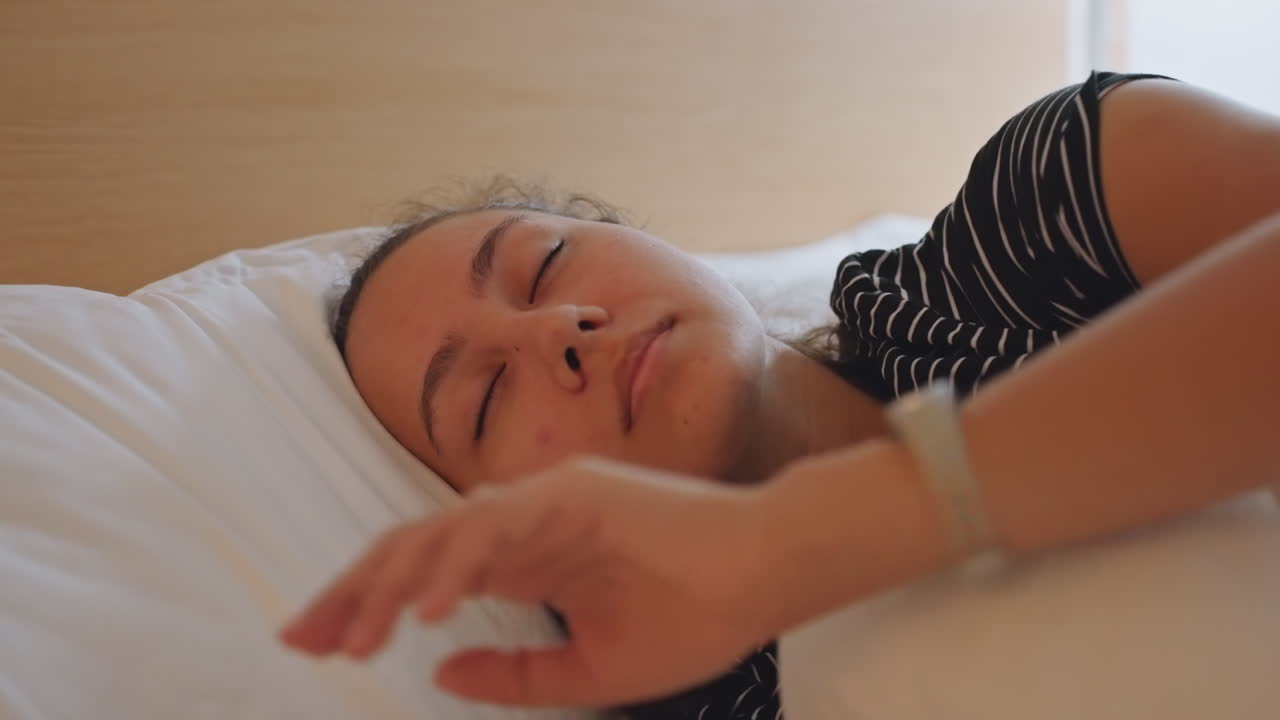 Hotelmorgen solopgang, ung kvinde ligger i sengen med blødt lys gennem gardinerne, strækker sig forsigtigt og blinker langsomt, mens gæsten vågner, hvid dyne og træhovedgærde indrammer en fredfyldt scene.