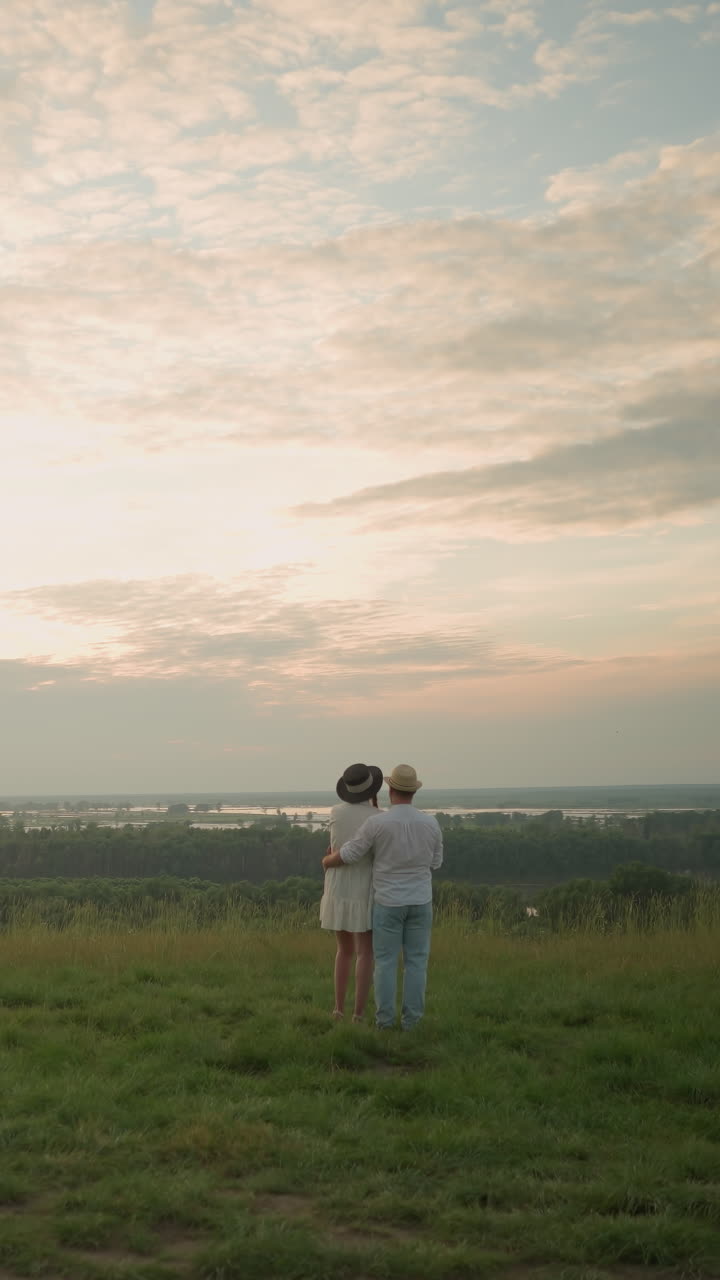 한 부부는 해가 지는 동안 호수 의 잔디 언덕 위에 서 있습니다. 남자는 색 셔츠, 모자, 청바지를 입고 여성의 허리를 잡고 있습니다. 그녀는 검은 모자와 색 드레스를 입고 있습니다.