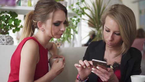 Two-young-girlfriends-using-smartphone-and-talking-in-cafe-and-drinking-coffee.-Shot-in-4k