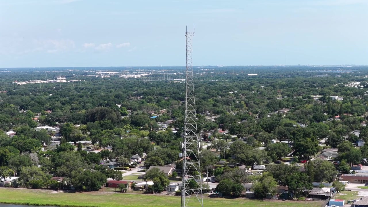 Alta torre di comunicazione si erge sopra un quartiere densamente alberato, un piccolo lago, tetti sparsi, uno skyline distante e sfocato, cielo soleggiato, Clearwater, Florida, Stati Uniti. Ampia ripresa aerea con drone.