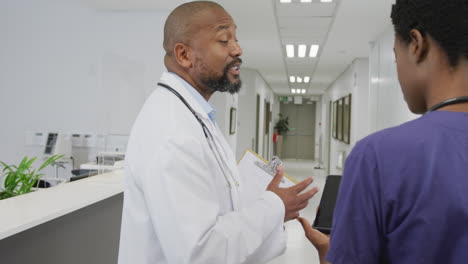 African-american-male-and-female-doctors-holding-clipboard-and-tablet,-talking-at-hospital