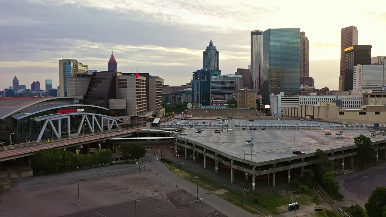 State Farm Arena with parking area and skyline of Atlanta Downtown during golden sunrise. Aerial approaching wide shot. Truist Plaza, Bank of America Plaza and Peachtree Tower with sunlight in back.