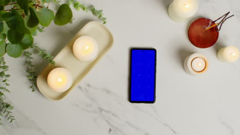 Overhead-View-Looking-Down-On-Still-Life-Of-Blue-Screen-Mobile-Phone-Lit-Candles-And-Incense-Stick-With-Green-Plant-And-Towels-As-Part-Of-Spa-Day-Decor-1