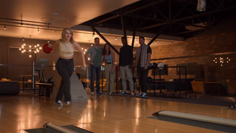 Eine-Junge-Frau-Beim-Bowling-Wirft-Einen-Ball-Auf-Die-Bahn,-Wirft-In-Zeitlupe-Einen-Ball-Ab-Und-Springt-Und-Tanzt-Vor-Freude.-Freunde-Fans-Unterstützen-Klatschen.-Eine-Gruppe-Multiethnischer-Freunde-Spielt-Gemeinsam-Bowling.