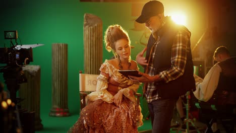 on period costume drama film set: beautiful smiling actress wearing renaissance dress sitting on a chair listens to movie director explaining to her scene context. high budget period drama. zoom in