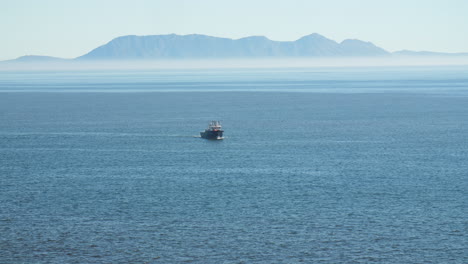 fishing trawler in distance off coast of south africa casting fishing nets as it turns in the water with heatwaves visible and misty blue mountains in background on sunny day
