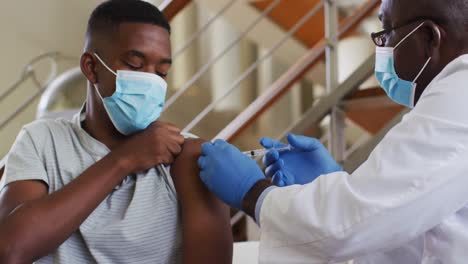 African-american-senior-male-doctor-giving-covid-vaccine-to-male-patient-in-home,-wearing-face-masks