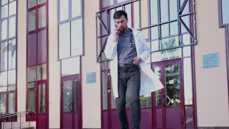 médico profesional con estetoscopio hablando en teléfono móvil saliendo frente al hospital. joven médico de sexo masculino con gafas y túnica blanca hablando en teléfono inteligente saliendo fuera de la clínica
