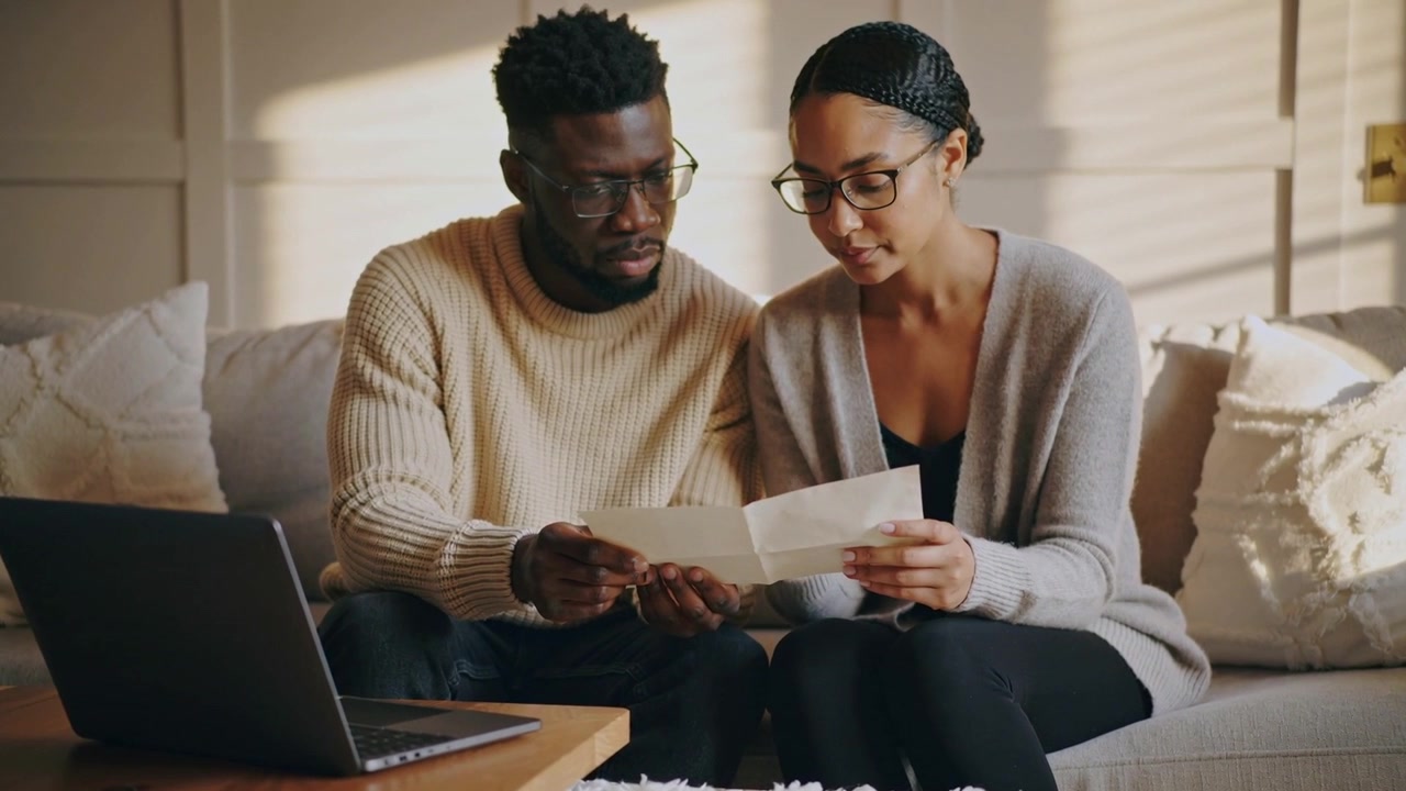 Holding folded paper, couple wearing sweaters leaning in, reading document on couch, laptop nearby