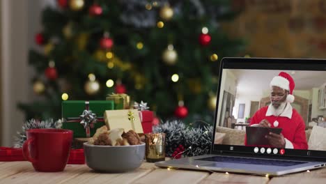 hombre afroamericano senior en disfraz de santa en videollamada en computadora portátil, con árbol de navidad