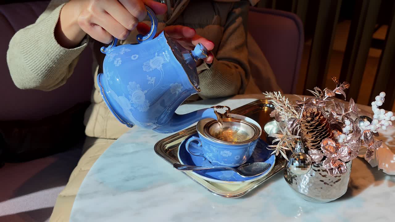 Close up of woman in beige puffer vest pouring hot yellow tea through silver strainer and drinking from blue ceramic cup at marble table in elegant lounge