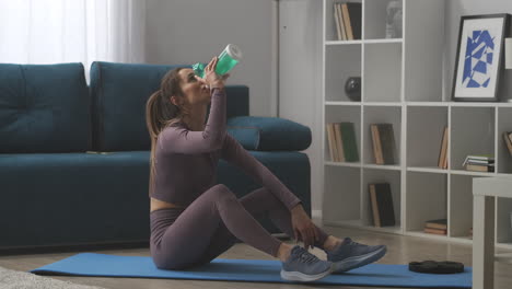 young sportswoman is drinking water at break during home fitness training sitting on floor in living room workout for body shaping