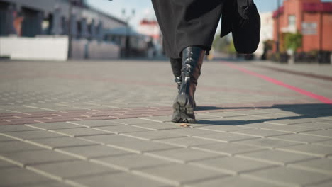 leg view of lady in black boots and coat holding a black briefcase strolling along an interlocked path in an urban setting, depicting a business-ready stride