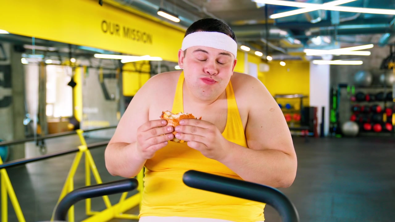 Uomo in sovrappeso che mangia un hamburger su una cyclette in palestra