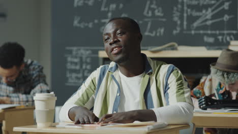 a young man sitting at a desk in a classroom with a blackboard in the background