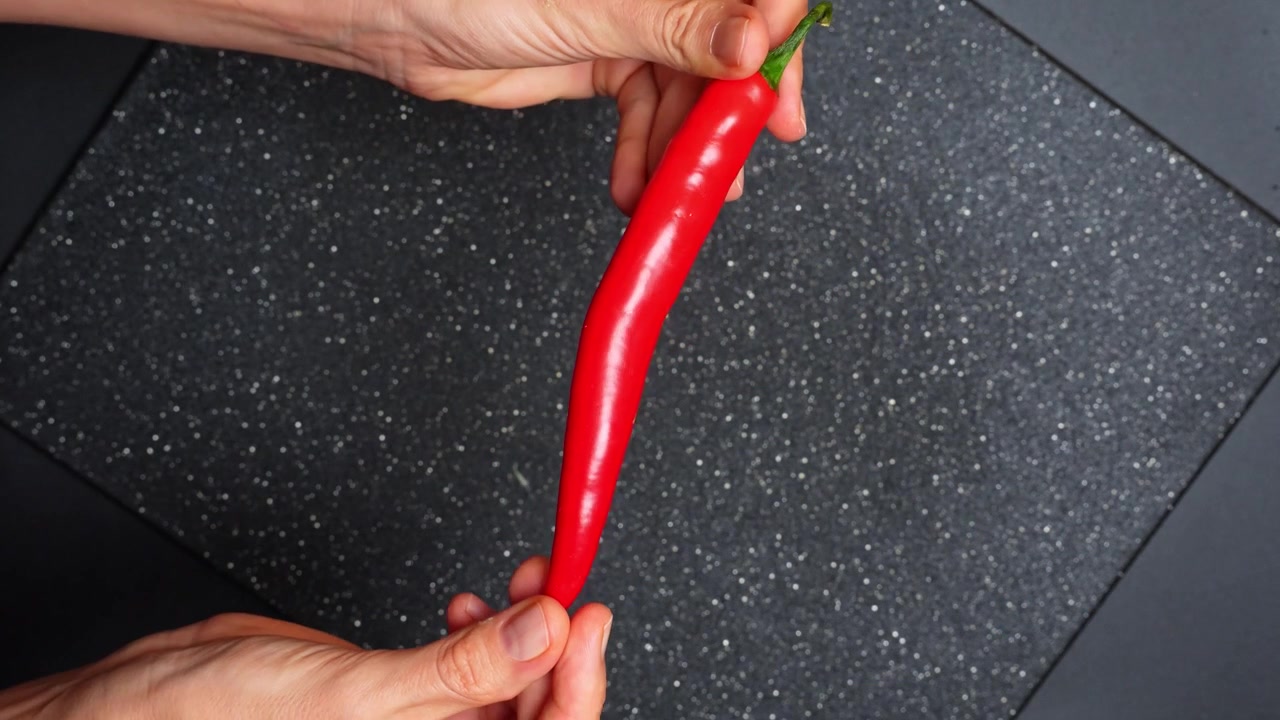 Mãos segurando uma pimenta vermelha inteira sobre uma tábua. Imagem aérea de uma pessoa exibindo uma pimenta vermelha longa e picante sobre uma tábua de corte preta salpicada