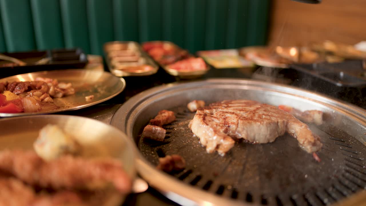 Hand flips pork steak with tongs on tabletop grill, warm lighting, shallow depth of field