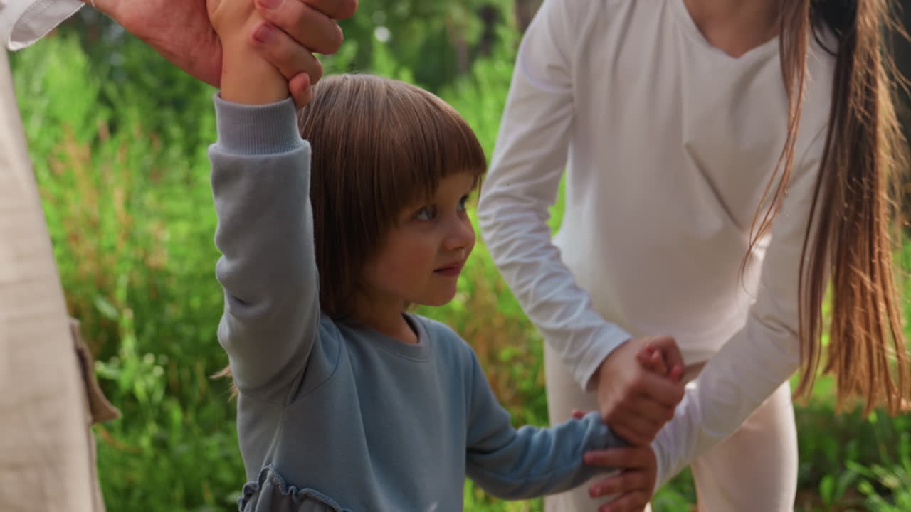 Lower view of brother and sisters walking on forest path holding hands, joyfully lifting youngest sibling between them, surrounded by lush greenery, creating playful outdoor family bonding moment