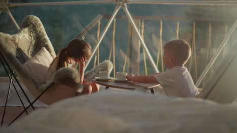 siblings rests sitting at table in hotel. boy pushes away empty cup at breakfast. brother and sister look at landscape in transparent glamping tent