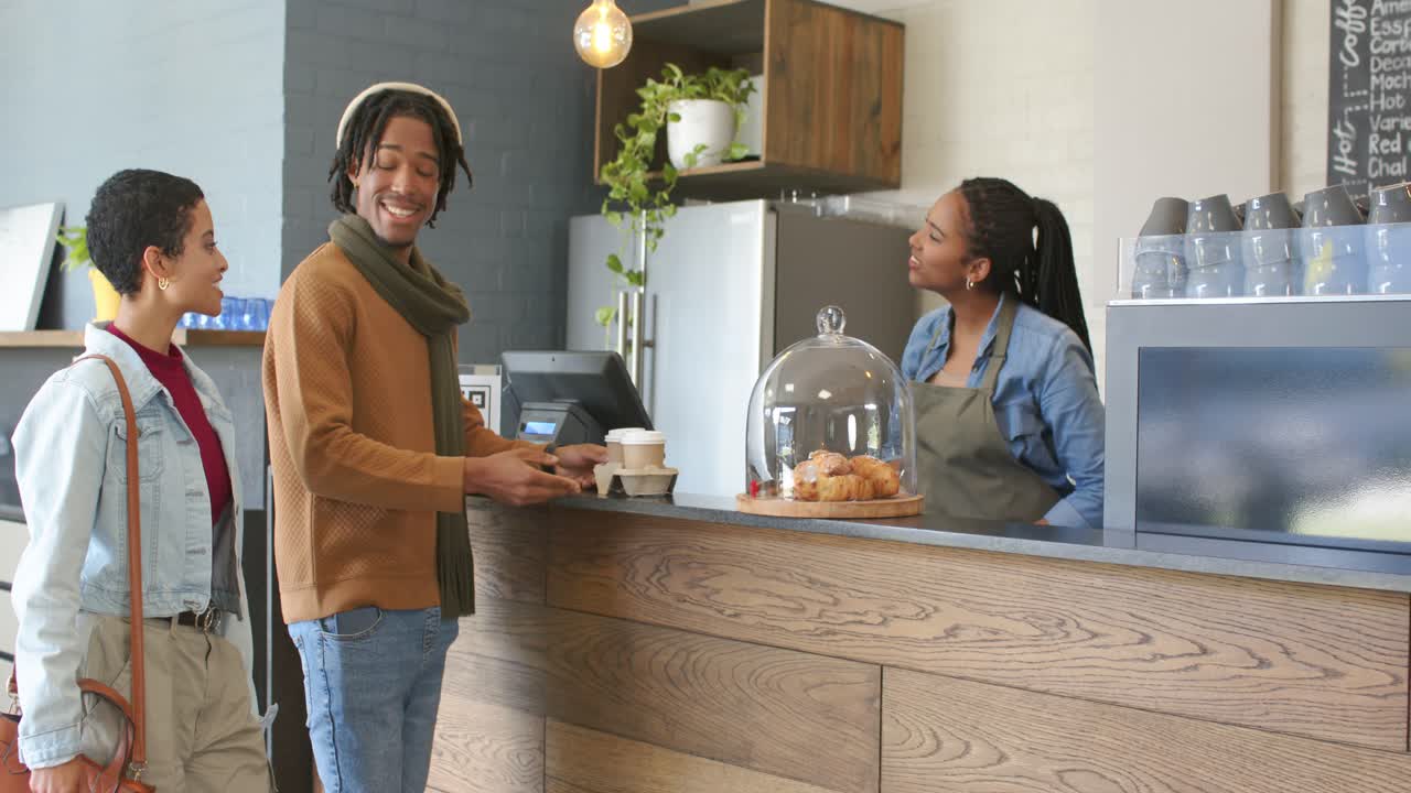 Verschiedenes Paar betritt ein Café, nachdem die Barista sie begrüßt hat, eine Bestellung aufgibt und Tassen abholt