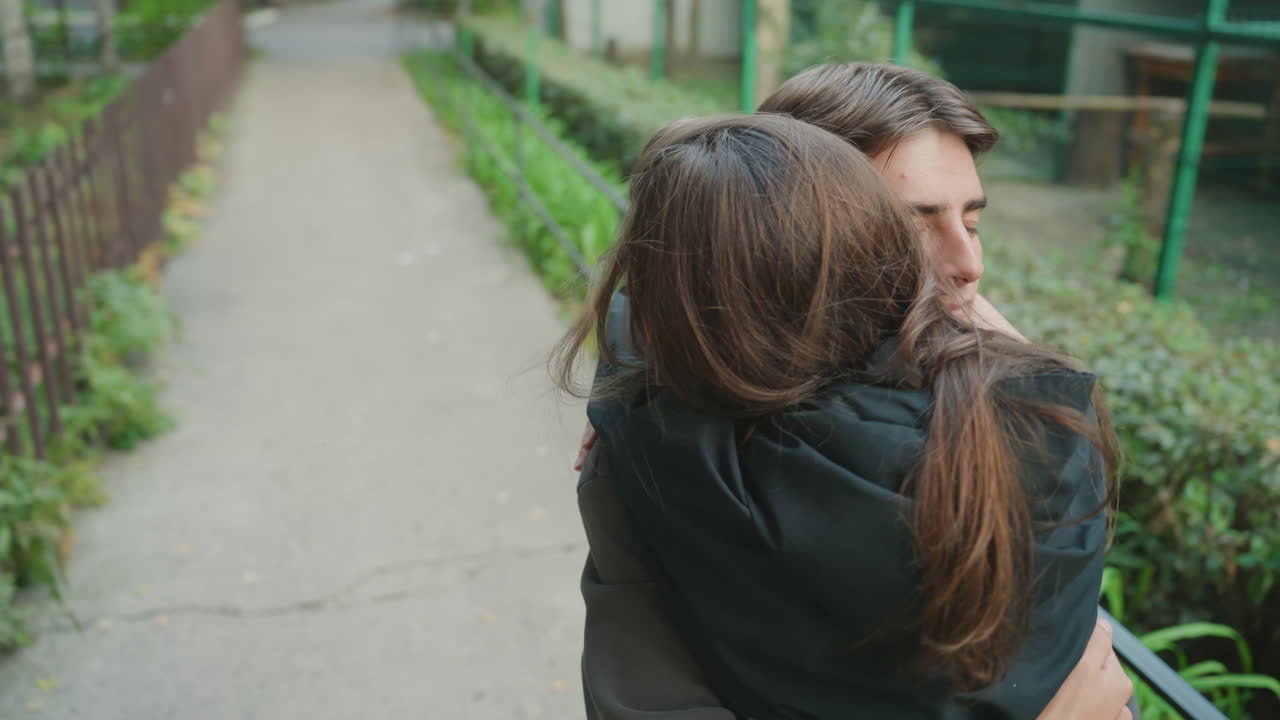 Young couple hugging on park path, emotional reunion and warm embrace between partners, smiling young man holding partner close near wooden fence and flower beds, natural light, candid handheld shot