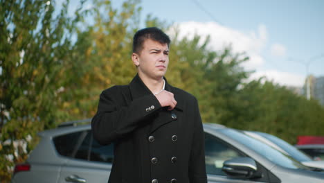 investor standing beside car adjusting collar confidently in outdoor setting with greenery and parked cars in background, projecting professionalism under clear sky in modern urban environment