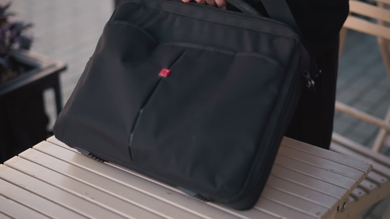 primer plano de una mujer con abrigo negro colocando una bolsa en una mesa de madera, abriéndola con los dedos adornados con anillos, sentada en un entorno al aire libre con flores borrosas y muebles en el fondo