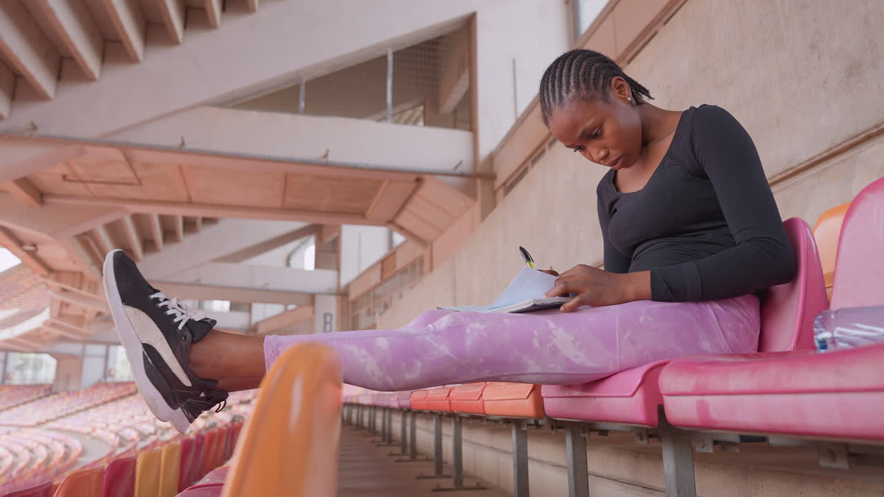 wide shot black woman eating snack on bleachers, pink seating and stadium architecture visible, sneakers and relaxed posture, casual spectator moment with warm tones and calm atmosphere