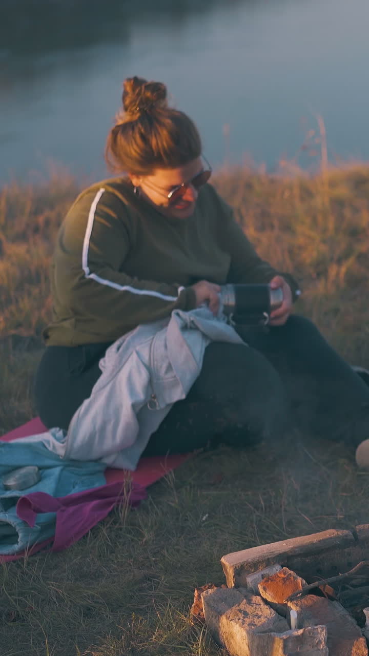 young man puts woods and twigs into bonfire and overweight girl sits on grass against river at bright sunset light