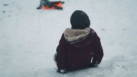 boy sitting on snowy hill slides down, slow motion view from behind