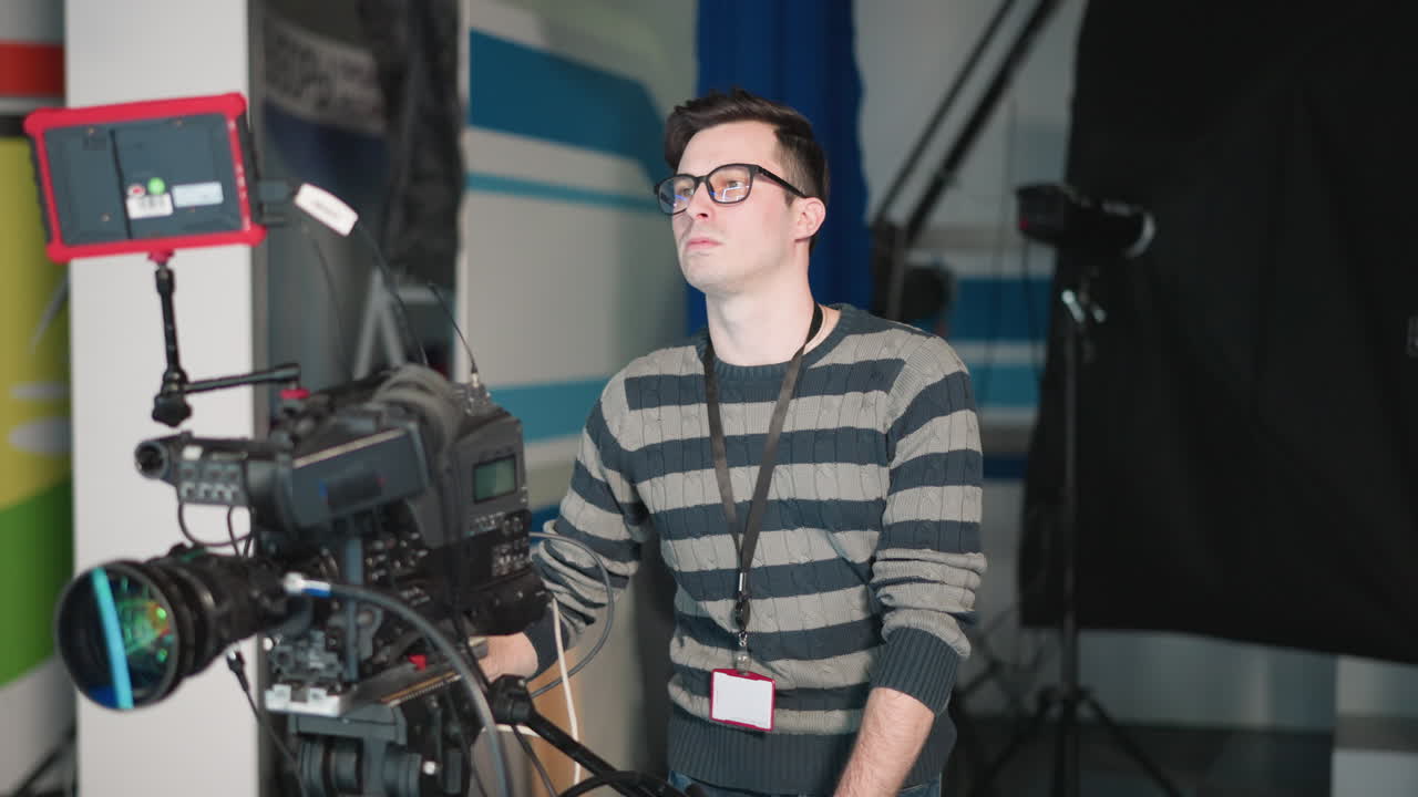 Close-up of camera operator adjusting broadcast camera with attached monitor in studio. Filming equipment and crew setup in modern TV production space