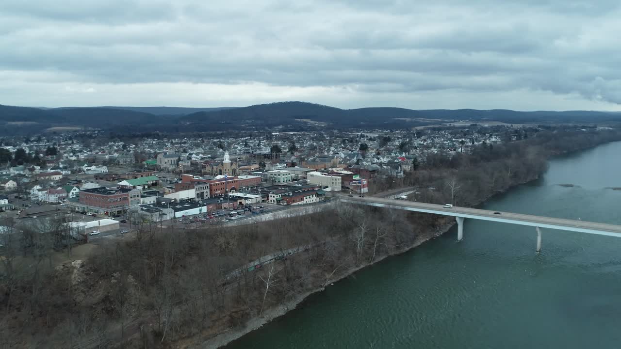 Una ripresa aerea in movimento sopra una fitta città di edifici, case, auto e negozi, che conduce verso un lago panoramico con un ponte che lo attraversa