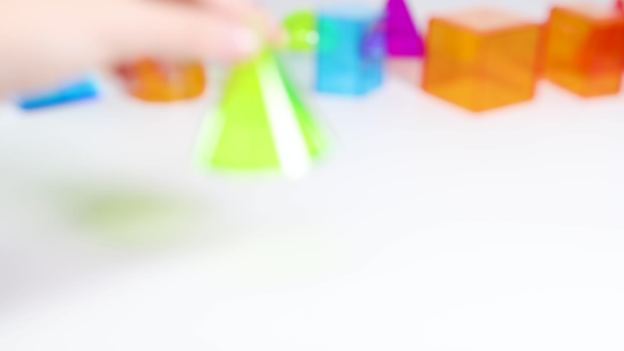 Hands manipulate vibrant 3D geometric shapes on a white surface, demonstrating movement and interaction in a bright setting