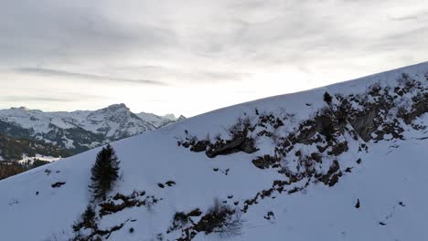 Aerial-revealing-flight-over-slope-of-mountains-showing-snowy-mountain-range-during-sunset-time-in-Amden,-Switzerland---Conifer-trees-in-the-valley