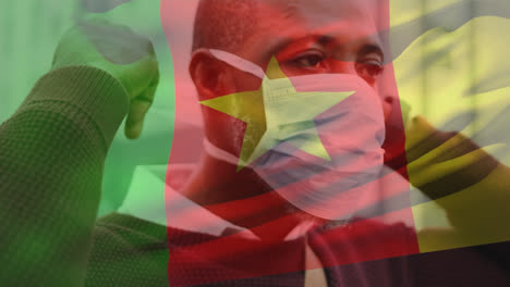 animation of flag of cameroon waving over man wearing face mask during covid 19 pandemic