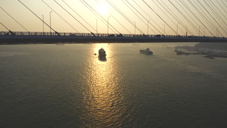 Aufsteigende-Aufnahme-über-Hängebrücke-Mit-Herannahendem-Frachtschiff-In-Der-Reflexion-Des-Sonnenlichts-Auf-Dem-Wasser