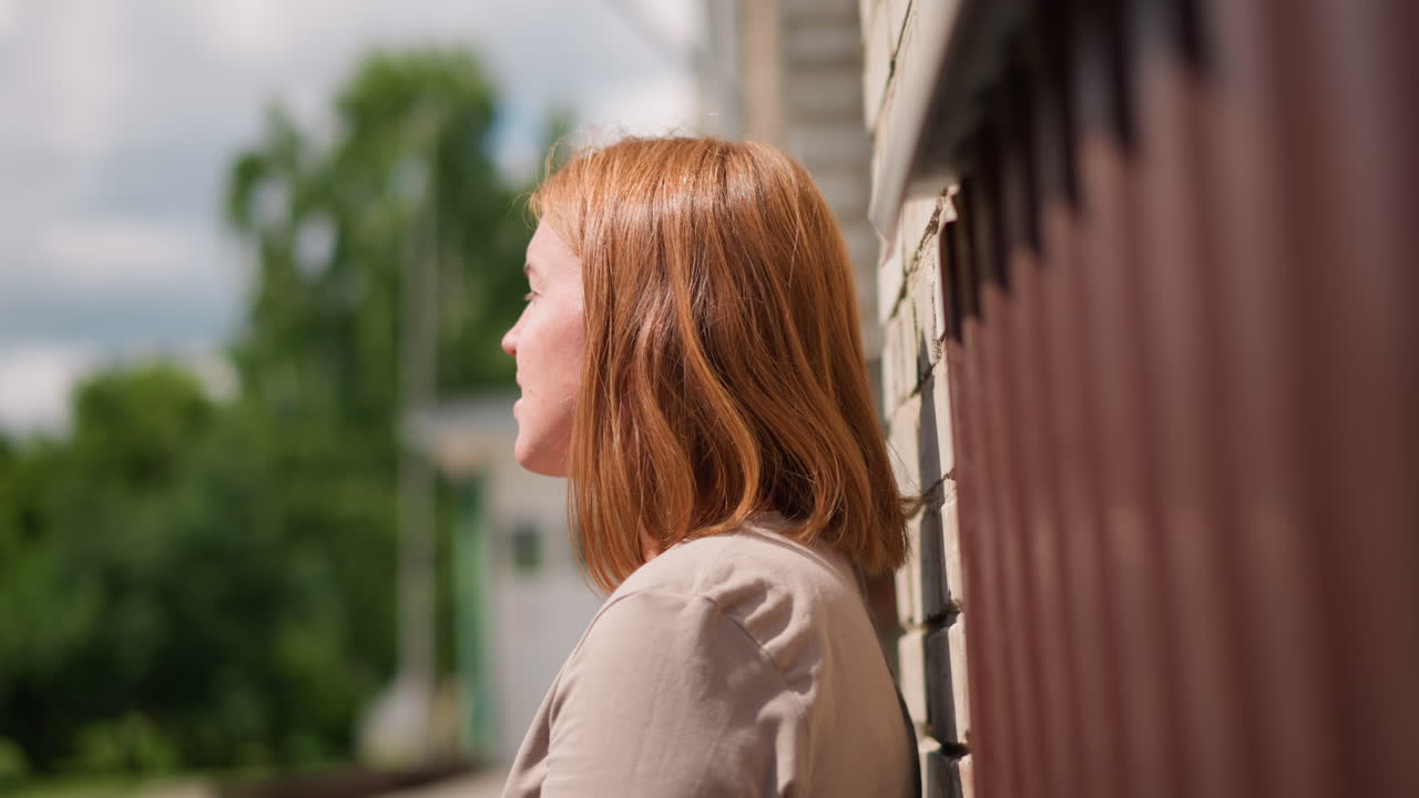 Worn out woman walking toward brick building under soft daylight, holding bag and pausing near wall, expressing fatigue, reflection, and emotional strain in peaceful outdoor scene