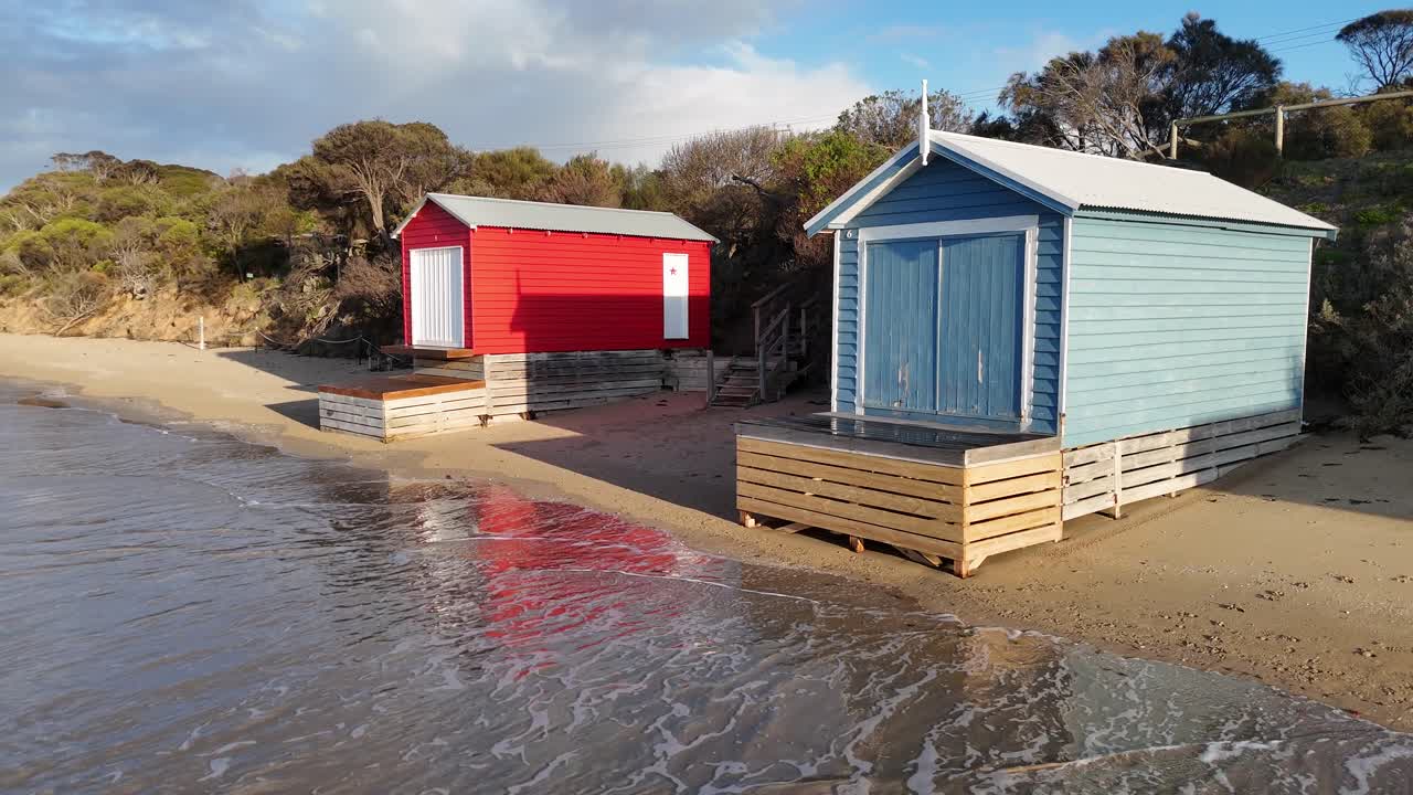 Gentle waves approach vibrant beach huts on sandy shore, morning sunlight, aerial camera movement
