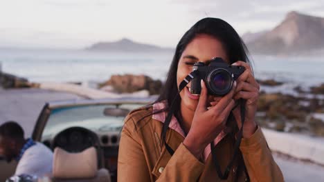 African-american-woman-using-digital-camera-to-take-pictures-while-standing-near-the-convertible-car