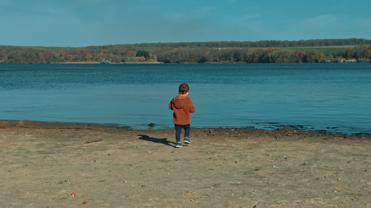 Rear view of a toddler in brown jacket walking to the river. Baby boy throws some stones into water and then turns to look for new ones.