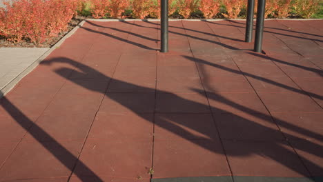 shadow silhouette of gym instructor performing side lunge on sunlit outdoor workout court, metal bars casting long shapes across rubber flooring bordered by autumn shrubs under clear sky