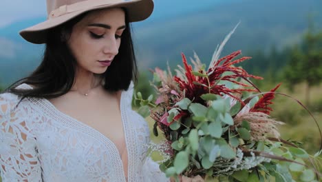 beautiful girl with wild flowers bouquet. young woman in hat looking at flowers in her hands on the background of mountains. close-up.