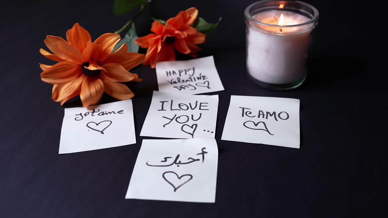 Fladt overblik over håndskrevne Valentinsdag-kærlighedsnoter på engelsk, fransk, spansk og arabisk med hjerter, varmt glas med stearinlys og orange blomster på mørk baggrund, romantisk hilsen med kopiområde