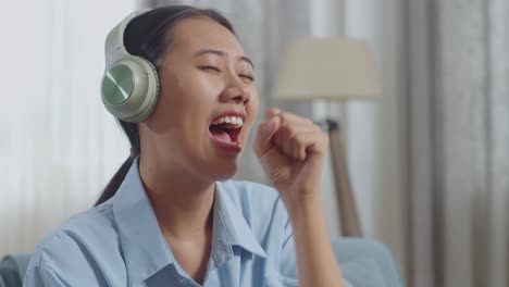 close up of asian woman with headphones listening to music and singing while sitting on sofa in the living room