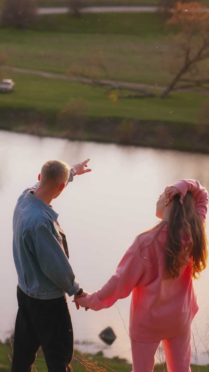 Young man and woman looking at the setting sun. Romantic couple holding hands while standing on the hill over the river at sunset. Rear view. Vertical video
