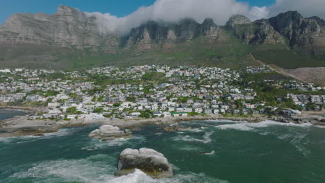 Houses-in-borough-in-slope-under-high-rocky-mountain-ridge.-Backwards-reveal-of-waves-crashing-on-coastal-rocks.-Cape-Town,-South-Africa