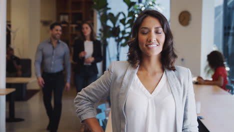 Portrait-Of-Businesswoman-Standing-In-Busy-Modern-Open-Plan-Office--With-Colleagues-In-Background