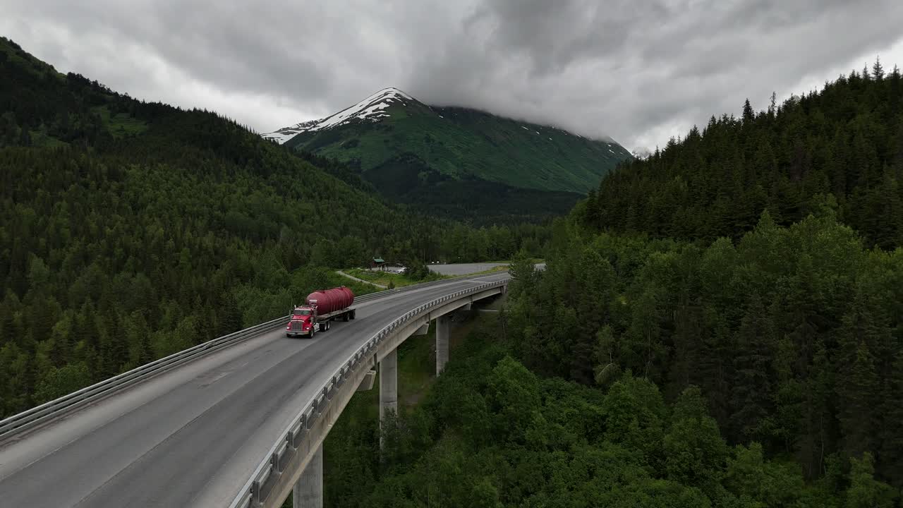 Cars and Semi Driving Over Bridge, Surrounded by Tall Sitka Spruce Trees on gloomy day, Mountain with a patch of snow on it, Canyon Creek Bridge in Alaska, Late June