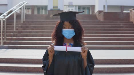 young african american female graduate standing in front of the camera with a diploma in her hands. the student has a protective medical mask. she stands on the stairs outside.